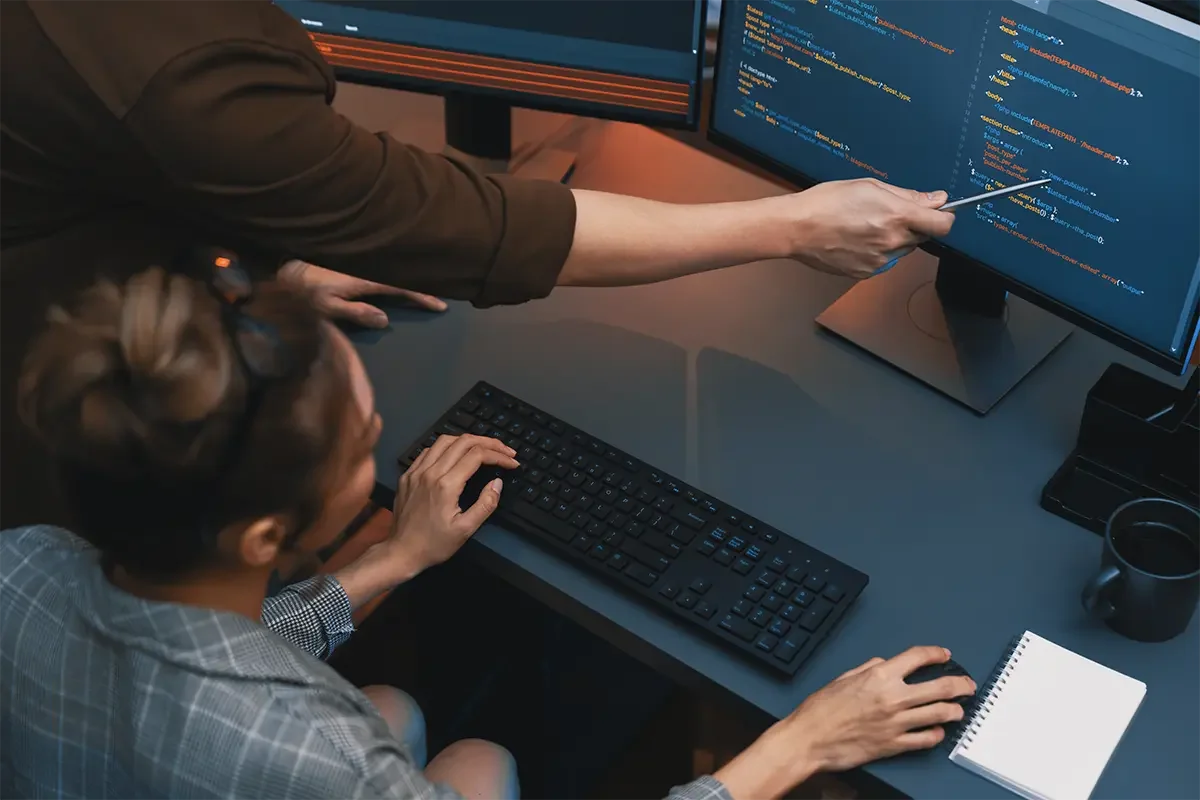 Dwoje programistów wspólnie analizujących kod na monitorach podczas tworzenia dedykowanej aplikacji — praca nad oprogramowaniem na zamówienie jako alternatywa dla gotowych rozwiązań SaaS